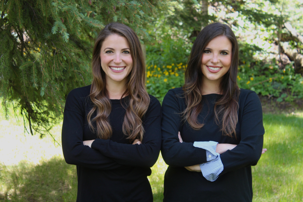 The co-founders of Dateability, Alexa and Jacqueline Child smiling in front of green trees and grass. They are both wearing black sweaters with their arms crossed.