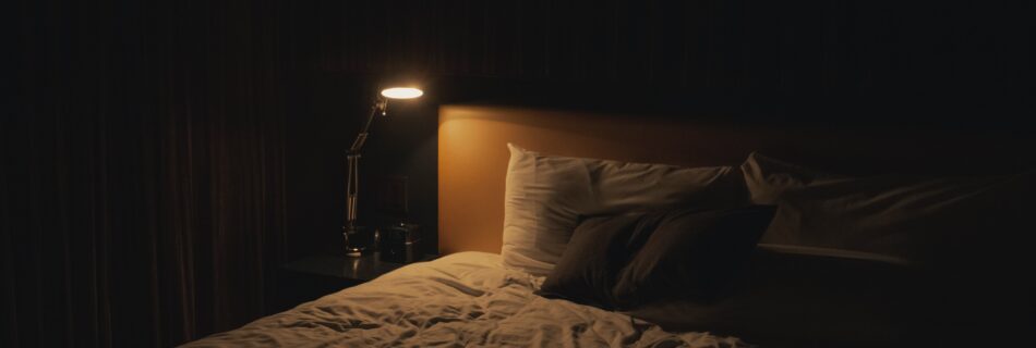 A dark room with a bed and a lamp shining on the nightstand.