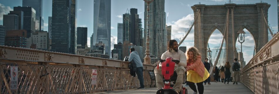 A still from the film showing a black male power wheelchair user being pushed by a white female over the Brooklyn Bridge.
