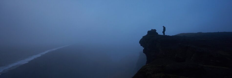 A dark and moody picture of a rock with a person at the edge of a cliff.