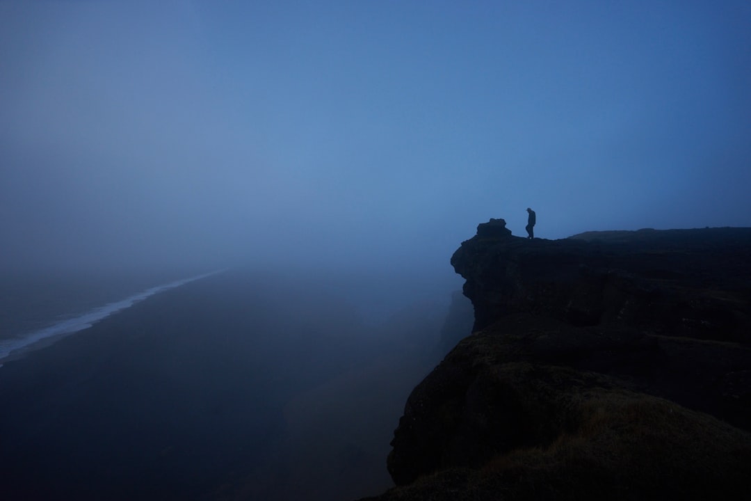 A dark and moody picture of a rock with a person at the edge of a cliff.