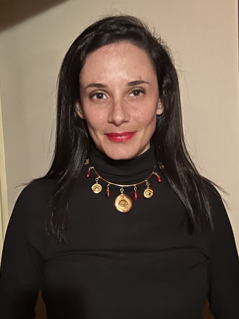 A white female woman with dark brown hair wearing a black turtleneck and a gold charm necklaces smiling softly.