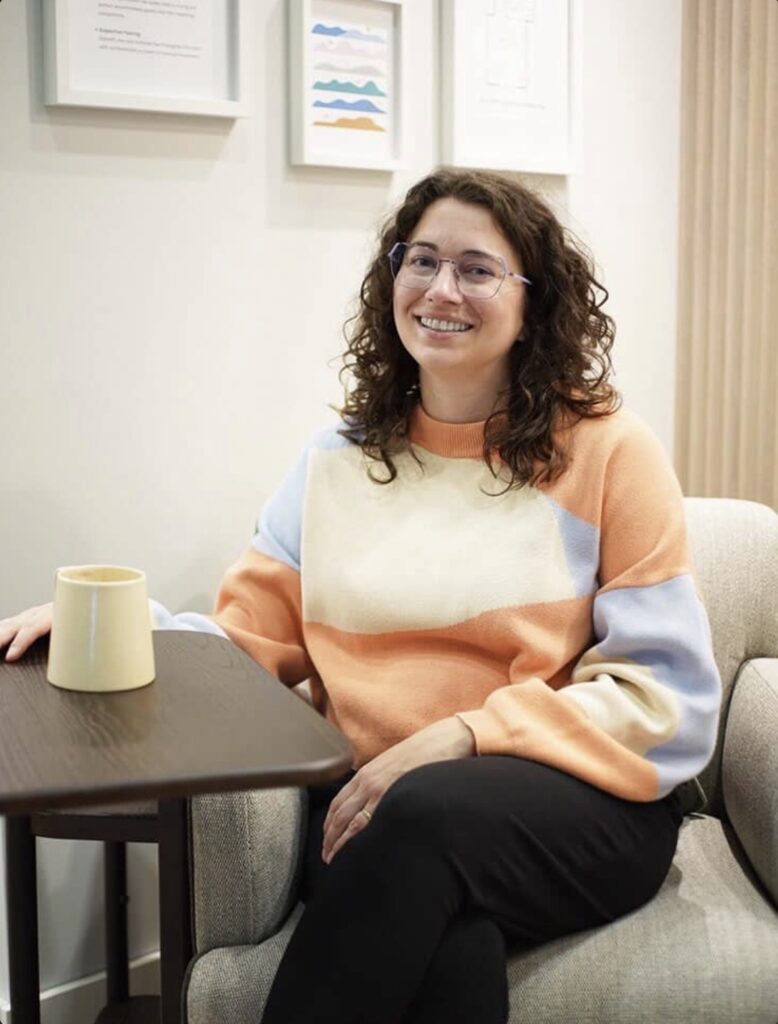 A white brunette woman smiling while sitting on a chair next to adaptive furniture.