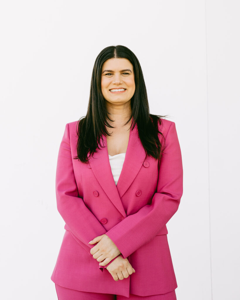 A white brunette woman wearing a hot pink pant suit smiling. 